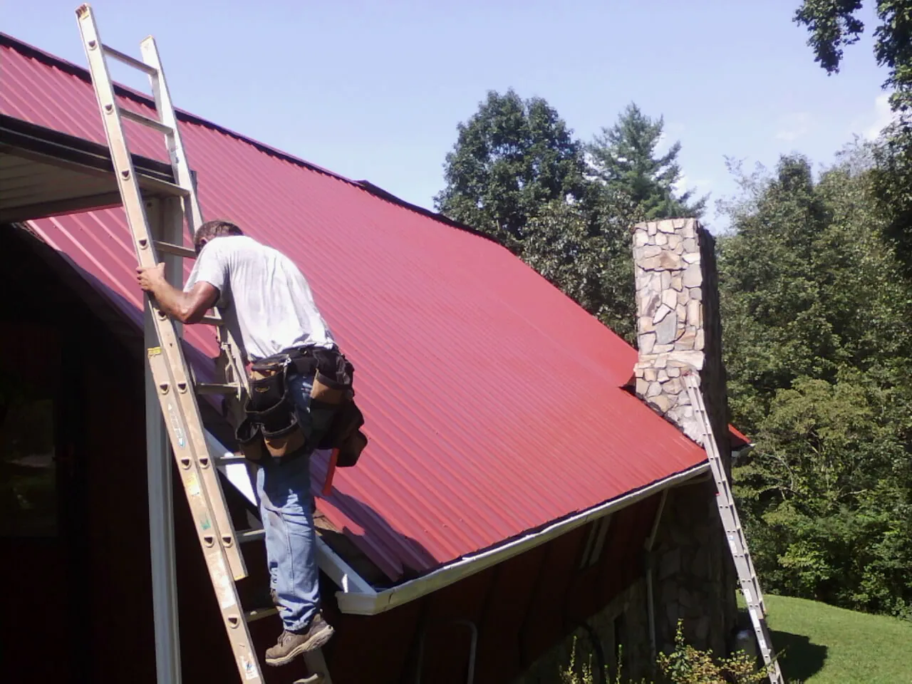 Roofer on a ladder beside a red metal roof and stone chimney — Soffit & Fascia Repair by Roofing Brothers in Doswell